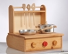 Petite cuisinière en bois avec 3 boutons, 1 rouge et 2 en bois naturel. Sur le dessus, 2 plaques de cuisson portant 1 poêle en aluminium avec manche en bois et une casserole avec poignées en bois et un bouton en bois sur  le couvercle. A l'arrière se trouve une parois en bois avec une étagère pour pendre les ustensiles qui en se refermant permet de porter la cuisinière et d'y ranger les jouets.