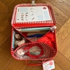 An open children's doctor's case shows its content: red hair protectnets, surgical masks, stethoscope, plastic syringes, paper compresses, wooden throat strips, blue plastic forceps, medical elastic bands,  plasters, wooden play thermometer, a wooden toy hammer,  printed medical notepad.