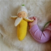 La main d'un tout petit bébé tient par un pan de sa pelure ouverte une banane poupée en tissu éponge jaune et blanc ayant un joli visage peint à la main.