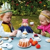 La poupée naturelle portant un bonnet rose et un gilet bleu marine est assise à table entre deux enfants qui jouent à la dinette au jardin.